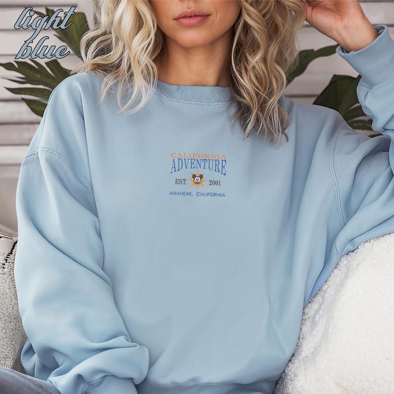 Pu&ograve; includere: Felpa azzurra con la scritta "CALIFORNIA ADVENTURE" in arancione e blu navy, con una piccola grafica di un personaggio dei cartoni animati. Sono visibili anche le scritte "EST. 2001" e "ANAHEIM, CALIFORNIA". Le parole "light blue" sono nell'angolo in alto a sinistra.