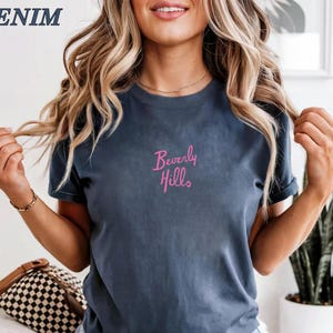 Peut inclure: Un t-shirt bleu denim avec le texte "Beverly Hills" en script rose. Le mot "DENIM" est dans le coin sup&eacute;rieur gauche. Le t-shirt est de style col rond.