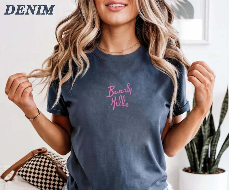 Peut inclure: Un t-shirt bleu denim avec le texte "Beverly Hills" en script rose. Le mot "DENIM" est dans le coin sup&eacute;rieur gauche. Le t-shirt est de style col rond.