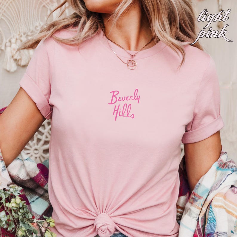 May include: Light pink t-shirt with the words "Beverly Hills" in a cursive font. The shirt has short sleeves and a knotted front. The words "light pink" are written in cursive in the upper right corner.
