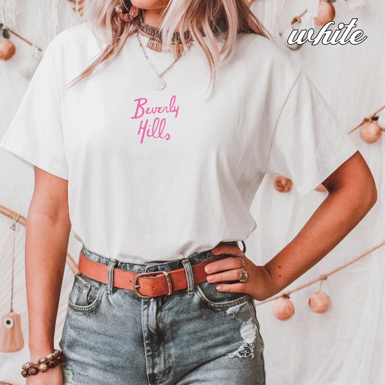 May include: White t-shirt with the text "Beverly Hills" in pink script. The person is wearing blue jeans and a brown belt. The word "white" is written in cursive in the upper right corner.