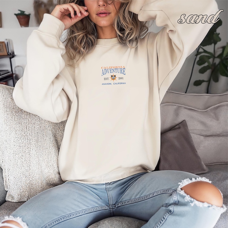 Pu&ograve; includere: Una felpa color sabbia con la scritta "CALIFORNIA ADVENTURE" in blu e arancione, con una piccola grafica e la scritta "EST. 2001 ANAHEIM, CALIFORNIA". La felpa &egrave; a maniche lunghe e ha un girocollo.