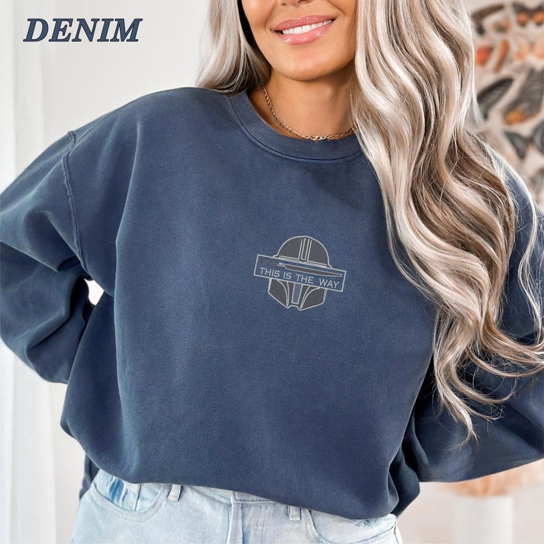 May include: Denim blue sweatshirt with a crew neck and long sleeves. The front features a gray graphic of a helmet and the text "THIS IS THE WAY" in a rectangular frame. The word "DENIM" is printed in the upper left corner.