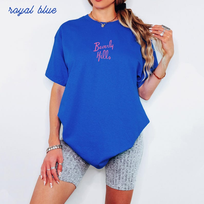 May include: Royal blue t-shirt with the text "Beverly Hills" in pink script. The shirt is a solid color and has short sleeves. The words "royal blue" are written in cursive in the upper left corner.