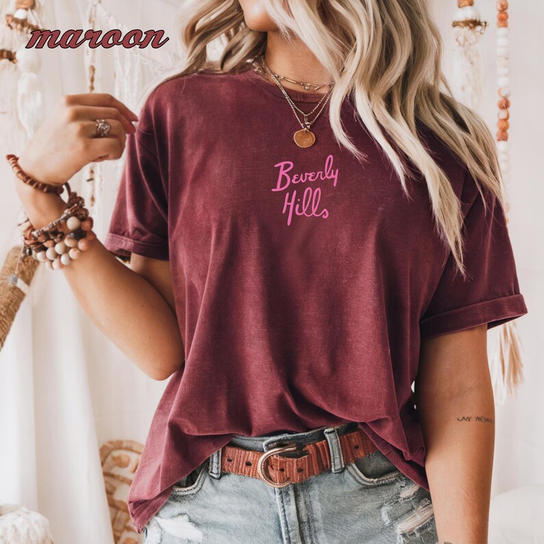 May include: Maroon t-shirt with the text "Beverly Hills" in pink script. The shirt has short sleeves and a crew neck. A brown belt with a gold buckle is visible. The model is wearing multiple bracelets and rings.