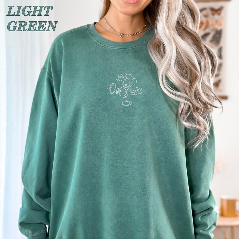 Op de afbeelding: Lichtgroene sweatshirt met een ronde hals en lange mouwen. De sweatshirt heeft een witte omtrek van een stripfiguur op de voorkant. De woorden "LIGHT GREEN" staan in de linkerbovenhoek gedrukt.