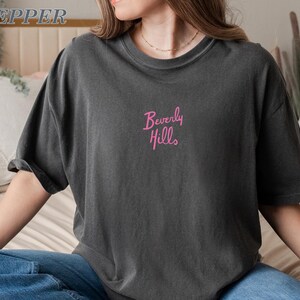 Peut inclure: T-shirt gris fonc&eacute; &agrave; manches courtes. Le texte "Beverly Hills" est brod&eacute; en rose sur la poitrine. Le t-shirt a une coupe d&eacute;contract&eacute;e et semble &ecirc;tre fait d'une mati&egrave;re douce.