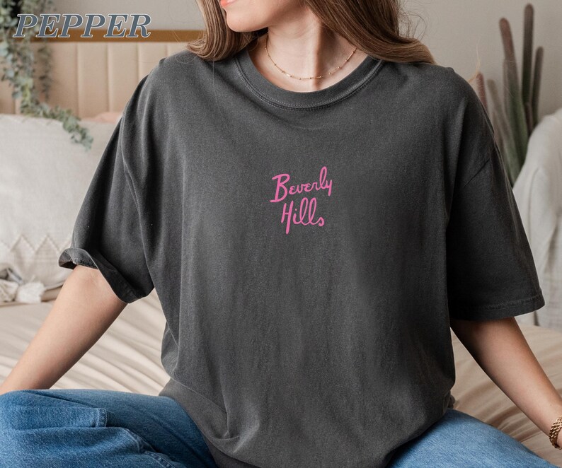 Peut inclure: T-shirt gris fonc&eacute; &agrave; manches courtes. Le texte "Beverly Hills" est brod&eacute; en rose sur la poitrine. Le t-shirt a une coupe d&eacute;contract&eacute;e et semble &ecirc;tre fait d'une mati&egrave;re douce.