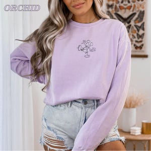 Op de afbeelding: Een lichtpaarse sweatshirt met een klein Mickey Mouse-omtrekdesign. Het woord "ORCHID" staat linksboven in het wit gedrukt. De top met lange mouwen wordt gecombineerd met een spijkerbroekshort.