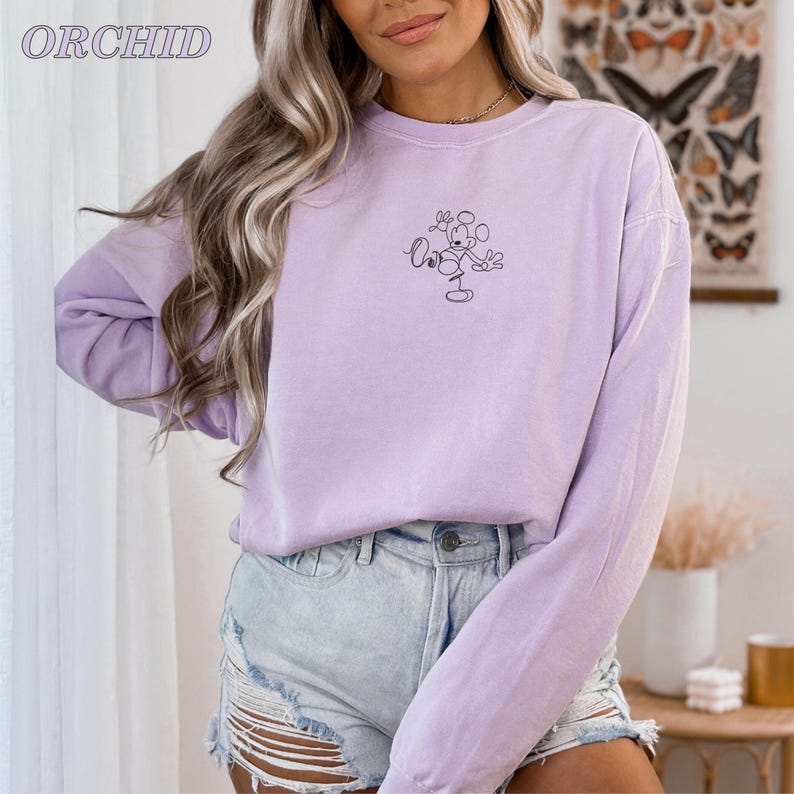 Op de afbeelding: Een lichtpaarse sweatshirt met een klein Mickey Mouse-omtrekdesign. Het woord "ORCHID" staat linksboven in het wit gedrukt. De top met lange mouwen wordt gecombineerd met een spijkerbroekshort.