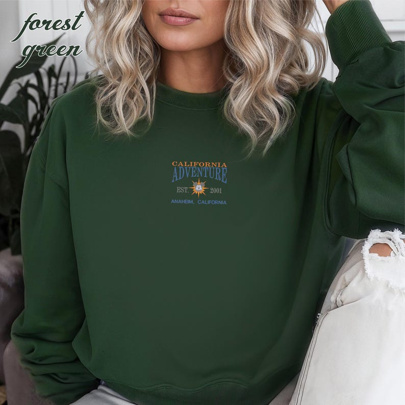 Pu&ograve; includere: Felpa verde foresta con la scritta "CALIFORNIA ADVENTURE" e un disegno a bussola. &Egrave; visibile anche il testo "EST. 2001" e "ANAHEIM, CALIFORNIA". La felpa &egrave; a tinta unita e ha maniche lunghe.