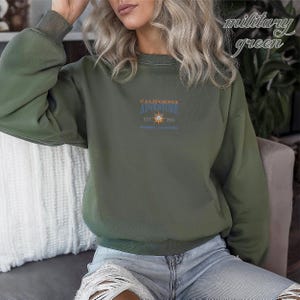 Pu&ograve; includere: Felpa verde militare con la scritta "CALIFORNIA ADVENTURE" e una grafica a forma di sole. La felpa ha un girocollo e maniche lunghe. Il testo "EST. 2001" e "ANAHEIM, CALIFORNIA" sono stampati anche sul davanti.
