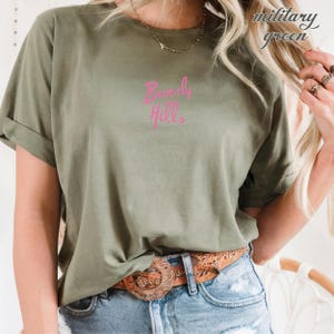 May include: Military green t-shirt with the text "Beverly Hills" in pink script. The shirt has rolled-up sleeves and is paired with blue jeans and a brown belt with a decorative buckle. A gold necklace is also visible.