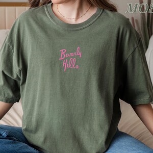 Peut inclure: Un t-shirt vert olive avec l'inscription "Beverly Hills" brod&eacute;e en rose sur la poitrine. Le t-shirt a des manches courtes et un col rond. Le style g&eacute;n&eacute;ral est d&eacute;contract&eacute; et ample.
