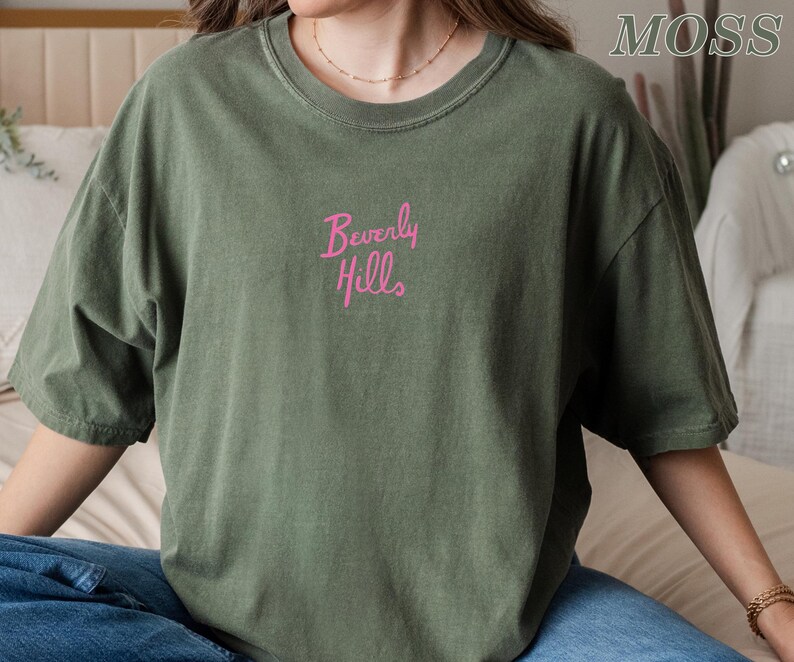 Peut inclure: Un t-shirt vert olive avec l'inscription "Beverly Hills" brod&eacute;e en rose sur la poitrine. Le t-shirt a des manches courtes et un col rond. Le style g&eacute;n&eacute;ral est d&eacute;contract&eacute; et ample.