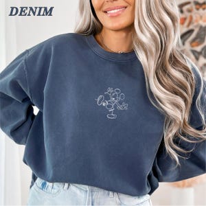 Op de afbeelding: Een denimblauwe sweatshirt met een wit geborduurd Mickey Mouse-ontwerp. Het woord "DENIM" is in het wit in de linkerbovenhoek gedrukt. De sweatshirt heeft een ronde hals en lange mouwen.