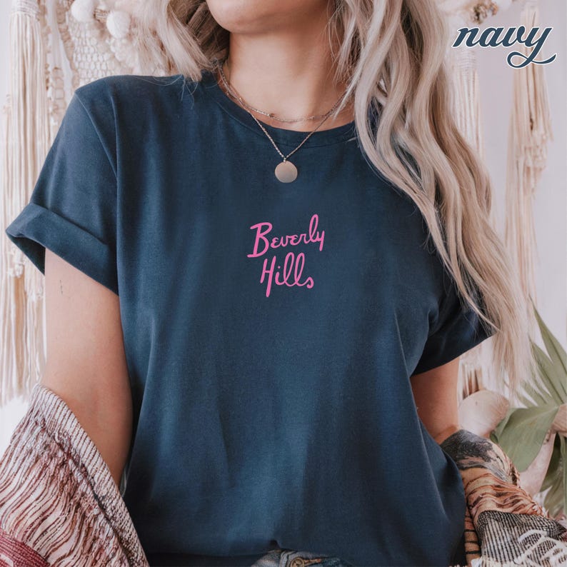 May include: Navy blue t-shirt with rolled sleeves. The shirt features the pink text "Beverly Hills" on the front. The word "navy" is written in cursive in the upper right corner.