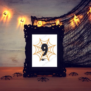 May include: A black framed picture with a white background and a black number nine inside an orange spider web. The picture is sitting on a wooden table with black plastic spiders scattered around it. There are string lights with orange jack-o-lantern shapes hanging in the background.