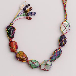 May include: A necklace featuring various colorful stones encased in a knotted net. The stones are in shades of red, green, brown, and purple, connected by a multicolored cord. The necklace has a loop at the top and a cluster of small stones.