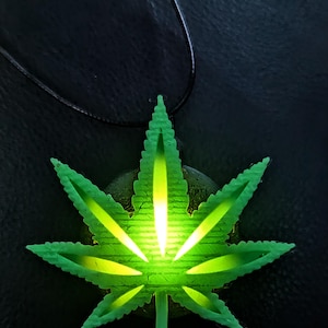May include: A green, glowing marijuana leaf pendant on a black cord. The pendant is a detailed, stylized representation of a cannabis leaf, with bright yellow-green illumination. The background is a dark, textured surface.