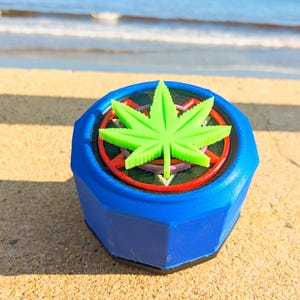 Könnte beinhalten: Ein blauer, achteckiger Grinder mit einem grünen Cannabisblatt oben. Der Grinder hat ein rotes und violettes Design um das Blatt herum. Das Produkt liegt auf einer sandigen Oberfläche mit dem Meer im Hintergrund.
