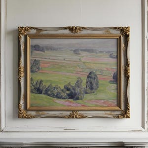 Puede incluir: Pintura al óleo de un paisaje con campos verdes y árboles, enmarcada en un marco decorativo dorado y gris. La obra de arte está montada en una pared blanca sobre una repisa gris.