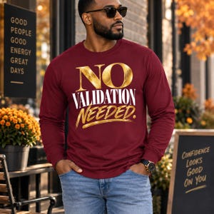 May include: A long-sleeve maroon crewneck sweatshirt featuring the text NO VALIDATION NEEDED in a combination of metallic gold and white lettering. The shirt is paired with blue denim jeans and a black wristwatch on the left wrist.