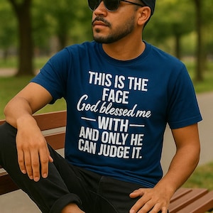 Könnte beinhalten: Marineblaues T-Shirt mit dem Text "THIS IS THE FACE God blessed me WITH - AND ONLY HE CAN JUDGE IT." Das Outfit umfasst eine schwarze Kappe, Sonnenbrille und eine schwarze Hose. Weiße Turnschuhe vervollständigen den Look.