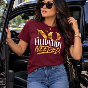 May include: A maroon short-sleeve t-shirt featuring the text NO VALIDATION NEEDED in a combination of metallic gold and white lettering. The shirt is paired with blue denim jeans that have a small distressed detail on the upper thigh area.