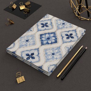 Puede incluir: Un cuaderno de tapa dura con un patrón repetitivo de motivos florales azules y blancos en una disposición de cuatrifolio. La cubierta muestra pétalos estilo acuarela en varios tonos de azul sobre un fondo blanco con finos bordes dorados.
