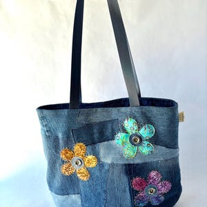 May include: A denim tote bag constructed from various shades of blue fabric patches. The bag features three fabric flower appliqués in yellow, teal, and purple, each centered with a metal button. It has two long dark blue shoulder straps.