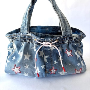 May include: A denim handbag featuring a light blue wash with a repeating white star pattern. The bag has two top handles, an elasticized opening, and a white drawstring cord with red-tipped ends. Some stars are outlined in red embroidery.