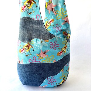 May include: A hobo-style shoulder bag constructed from alternating panels of dark blue denim fabric and a light blue cotton print featuring Spongebob Squarepants and Patrick Star characters alongside pink and yellow flower shapes.