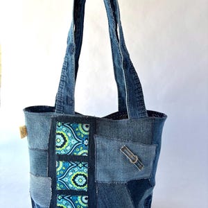 May include: A patchwork tote bag constructed from various shades of blue denim fabric. The bag features a vertical panel with a bright blue, green, and black paisley pattern, two denim shoulder straps, and a small metal buckle detail on the front.