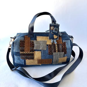 May include: A barrel-shaped handbag constructed from blue denim and various textured fabric patches in shades of brown, tan, and gray. The bag features black nylon handles and a detachable shoulder strap, with a small denim tag attached.