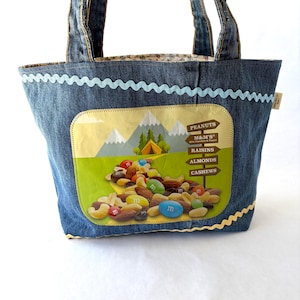 May include: A denim tote bag featuring a rectangular graphic of a mountain landscape with a tent and a trail of mixed nuts and M&M's candies. A wooden signpost lists Peanuts, M&M's, Raisins, Almonds, and Cashews. The bag has light blue rickrack trim.