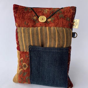 May include: A patchwork bag with a denim pocket, featuring a button closure and a black elastic loop. The bag incorporates fabrics in shades of red, gold, and blue, with a striped pattern and floral accents. A small tag is attached.