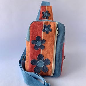 May include: A blue denim sling bag with a striped orange and red front panel. The bag features three denim flower appliqués and a blue adjustable strap. A white zipper runs down the side.