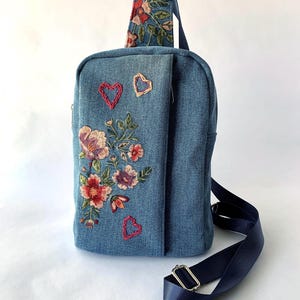 May include: A denim sling bag with floral embroidery in red, pink, and purple. The bag features two embroidered hearts and a navy blue adjustable strap. The bag's design includes a small pocket with floral embroidery.
