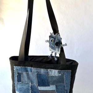 May include: A black tote bag featuring a rectangular patch made of various blue denim fabric scraps in different shades. A small, handmade denim robot ornament with button eyes and stitched details is attached to one of the black nylon shoulder straps.