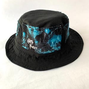 May include: Black bucket hat with a blue and white graphic featuring the text "Harry Potter" and a castle design. The hat has a wide brim and a distressed, frayed edge on the graphic panel.