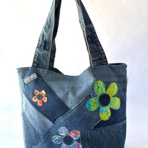 May include: A tote bag constructed from various shades of blue denim fabric in a patchwork design. The bag features two denim handles and three fabric flower appliqués in yellow, green, pink, and blue patterns with dark circular centres.