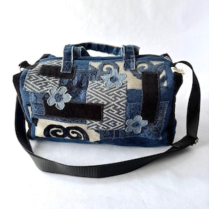 May include: A patchwork denim bag with a black adjustable shoulder strap and two handles. The bag features various blue denim patches with floral and geometric patterns. Decorative blue flower appliqués are sewn onto the bag.