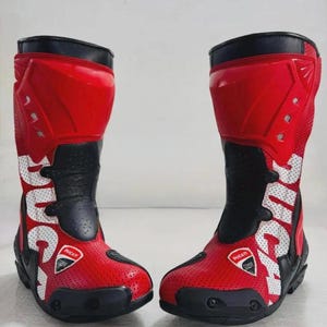 Puede incluir: Par de botas de moto rojas y negras con la palabra "DUCATI" en letras blancas. Las botas tienen un diseño perforado en las secciones rojas y presentan el logotipo de Ducati. Las botas están diseñadas para protección y estilo.