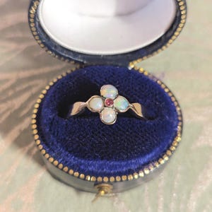 May include: A silver ring with a four-petal design, each petal featuring an opal stone. A small, round red stone is set in the centre. The ring is displayed in a blue velvet jewellery box with a gold-coloured trim.