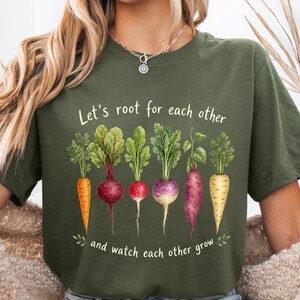 Puede incluir: Camiseta verde oliva con una ilustración de acuarela de varias hortalizas de raíz, incluyendo zanahorias, remolachas y nabos. El texto "Let's root for each other and watch each other grow" está impreso encima.