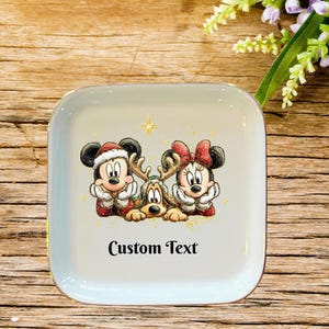 May include: A square white ceramic trinket dish featuring a festive illustration of Mickey Mouse in a Santa hat, Minnie Mouse with a red bow, and Pluto wearing reindeer antlers. The text Custom Text is printed in black below the characters.
