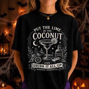 May include: A black t-shirt featuring a white graphic print of a margarita glass, a lime, and a bottle labeled Witches Spirits. The text reads Put the lime in the coconut and drink it all up, with Midnight Margaritas at the bottom of the design.