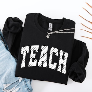 Könnte beinhalten: Ein schwarzes Langarm-Sweatshirt mit Rundhalsausschnitt, das das Wort TEACH in großen, weißen Blockbuchstaben mit schwarzem Punktmuster zeigt. Das Kleidungsstück hat ein Gildan-Markenetikett am inneren Halsausschnitt und eine blaue Jeans.