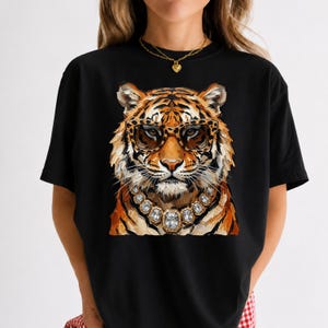 May include: A black short-sleeved t-shirt featuring a graphic print of a tiger wearing leopard-print glasses and a chunky silver-toned necklace with large clear rectangular crystals. The tiger has orange and black fur with white whiskers.